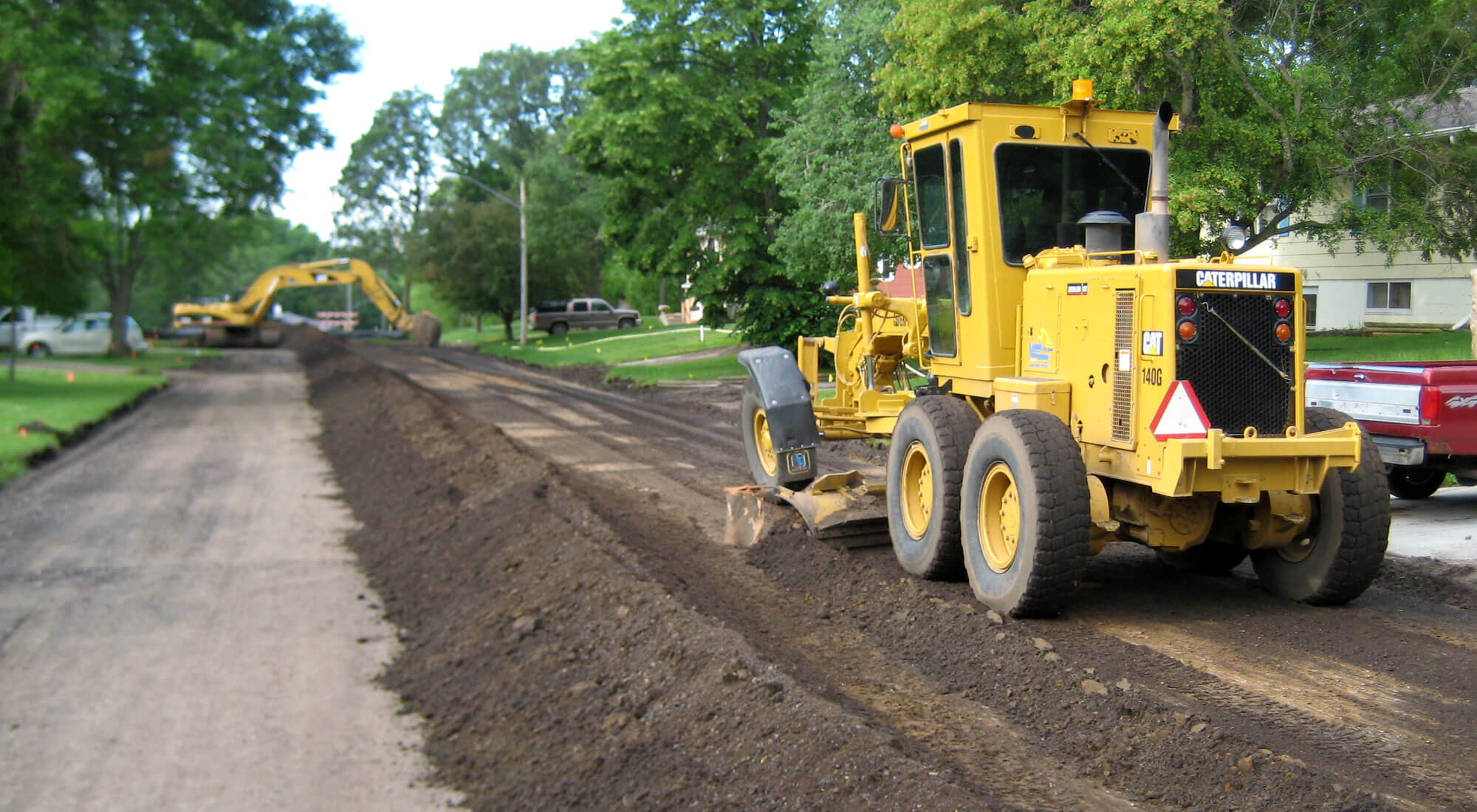 Road grader in the foreground smoothing a fresh layer of aggregate with an excavator in the background